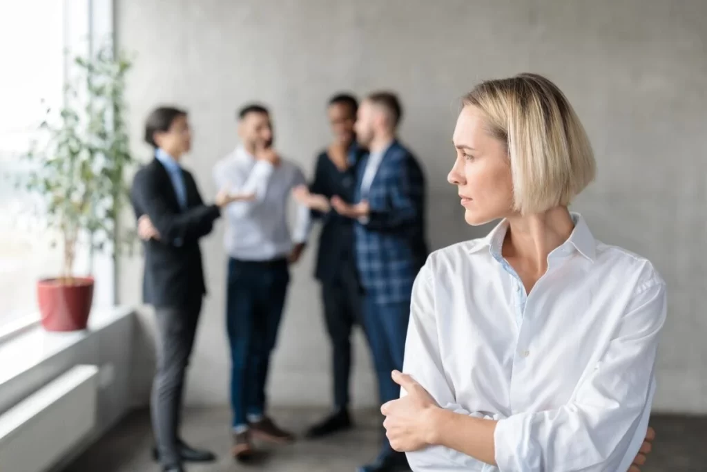 Male Coworkers Whispering Behind Back Of Unhappy Businesswoman In Office E1618831419720