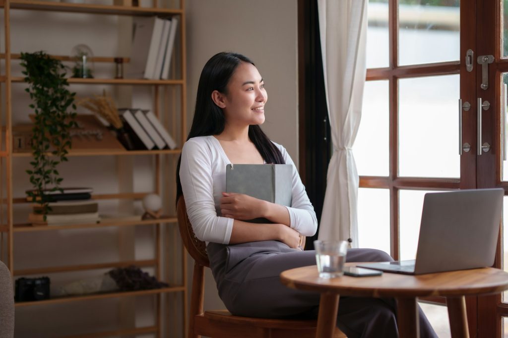 Happy Asian Female College Student Reading Book And Working On Her Laptop In The Living Room During