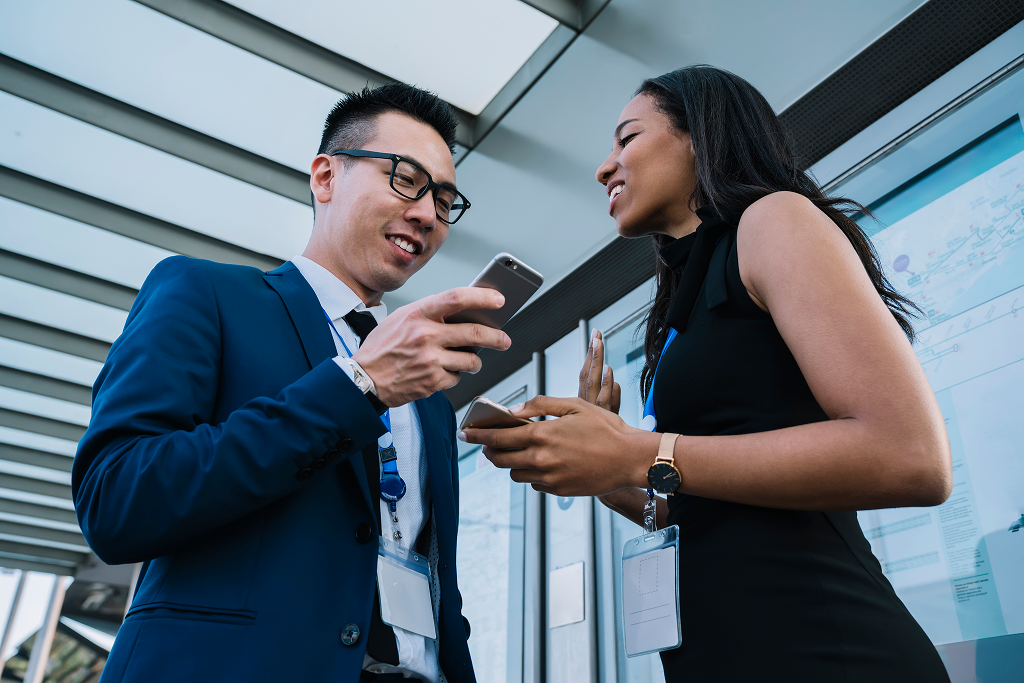 Employee Engaging In Workplace Communication Standing Next To A Colleague While Using A Cellphone