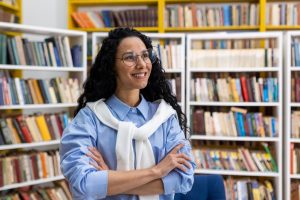 Confident Female Librarian Standing In Library Bookshelves